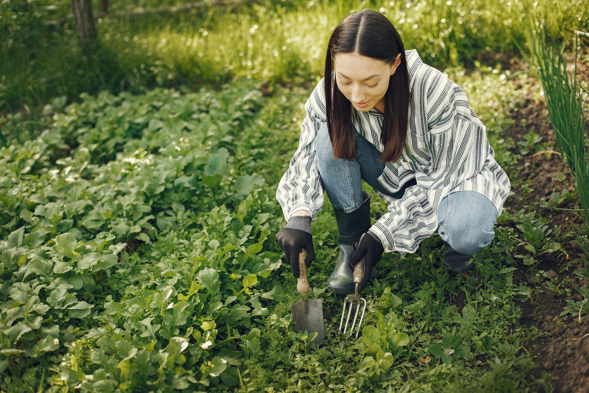 A young woman tends to her garden, cultivating vibrant greens with garden tools on a sunny day.